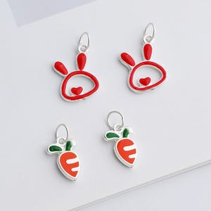 May include: Four silver charms with red and white enamel designs. Two charms are bunny-shaped with red ears and a heart, and two are carrot-shaped with green tops and orange bodies. Each charm has a small loop for attaching to jewelry.