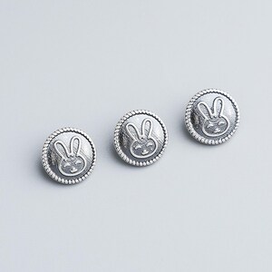May include: Three silver-colored buttons with a raised bunny face design. Each button has a rope-like border. The bunny's face is smiling, with long ears and a round face. The buttons are on a light gray background.