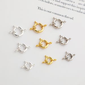 May include: A selection of circular jewellery clasps in gold and silver. Each clasp features a ring design with small bars extending outwards. The background is a white surface with text.