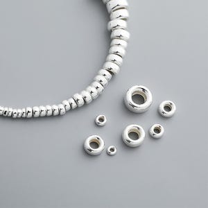 May include: A selection of shiny silver beads in different sizes. Some are strung together, forming a curved line, while others are loose. The beads have a smooth, reflective surface, and are displayed on a grey background.