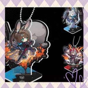 May include: Acrylic character standees with anime-style illustrations. The stands show various characters with detailed designs, including one with bunny ears and a sword. The stands have a base for display and a chain for hanging.