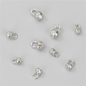 May include: A collection of small, silver-colored jewelry clasps scattered on a light gray surface. Each clasp has a rounded, barrel-shaped body with a loop on one end and a curved opening on the other, designed for securing jewelry.