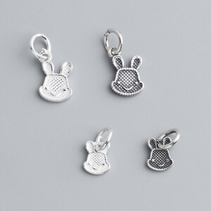 May include: Four small silver-colored bunny charms with a textured surface and a loop for attaching to a bracelet or necklace. The charms are displayed against a gray background.