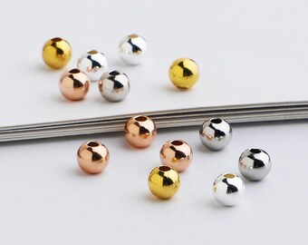 May include: A collection of small, round beads in various metallic finishes: gold, rose gold, and silver. The beads are scattered across a white surface, showcasing their reflective qualities and smooth texture. Ideal for jewelry making and crafting.