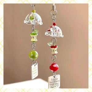 May include: Two decorative hanging ornaments. Each features a clear umbrella-shaped top, apple-shaped beads, and a small metal tag. The tags read "follow your dreams, trust your heart". One ornament has a green apple bead, the other a red one.