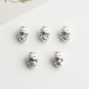 May include: Five small, silver-colored charms with a smiling, cartoon-like face. Each charm has a different design on its belly, including a coin, a symbol, and a wrapped bundle. The charms are arranged on a white surface.