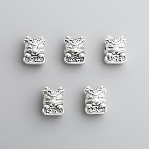 May include: Five silver-colored charm beads in the shape of smiling cartoon tigers. Each bead has detailed facial features and Chinese characters. The beads are displayed against a plain, light gray background.