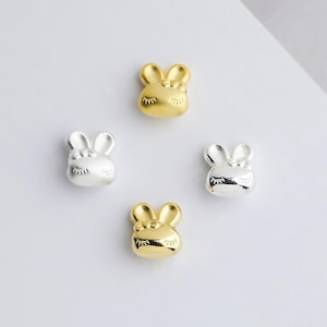 May include: Four small, bunny-shaped charms in gold and silver. Each charm features a sleeping face with closed eyes and long eyelashes. The charms are arranged on a white background.