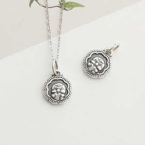 May include: A silver-colored pendant necklace and a matching charm. The pendant and charm feature a detailed, embossed design of a small creature within a scalloped border. The necklace chain is delicate and silver. The background is white.