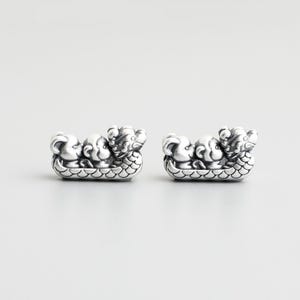 May include: Two silver-colored dragon boat charms. Each charm features a boat with three stylized dragon figures. The boats have a scale-like pattern on the bottom. The background is a plain, light gray.