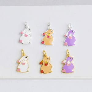 May include: Six colorful bunny charms in white, tan, and purple with pink and red accents. Each charm has a silver or gold-colored bail for attaching to a necklace or bracelet. The charms are arranged on a white surface.
