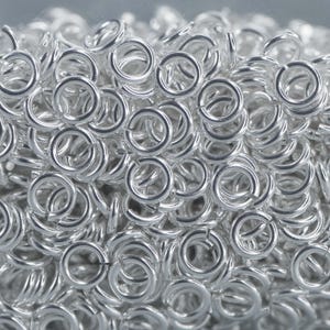 Puede incluir: Una imagen de primer plano de un montón de anillos de salto de plata. Los anillos son pequeños y redondos, y todos son del mismo tamaño. Son perfectos para la elaboración de joyas u otros proyectos de artesanía.