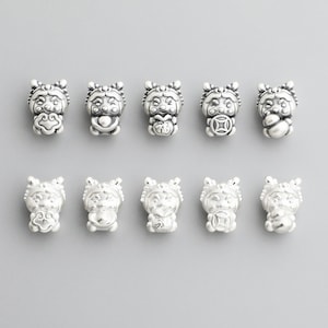 May include: Ten small, silver-colored charm beads in a row. Each bead is a detailed, miniature figure with a smiling face, ornate headdress, and a round body. The background is a solid, light gray.
