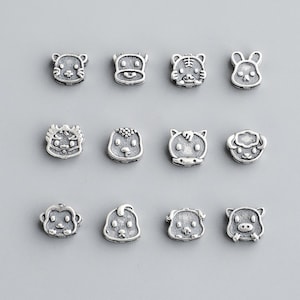 May include: A collection of twelve small, silver-colored charms, each depicting a different animal face. The charms are arranged in three rows of four, with a variety of animals including a tiger, rabbit, and pig. The charms have a detailed, slightly aged appearance.