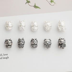 May include: A collection of ten small, silver-colored charm beads. Each bead is sculpted in the shape of a stylized animal, possibly a tiger or lion, with intricate details. Some beads appear to hold objects. The beads are arranged on a white surface.