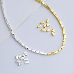 Peut inclure: Un collier en argent et or composé de petites perles rectangulaires. Le collier est disposé sur une surface blanche avec des perles supplémentaires éparpillées autour.