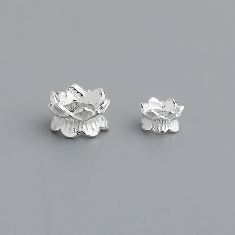 Puede incluir: Dos abalorios de plata con forma de flor de loto, uno m&aacute;s grande que el otro. Los abalorios son detallados y tienen un aspecto delicado y femenino.