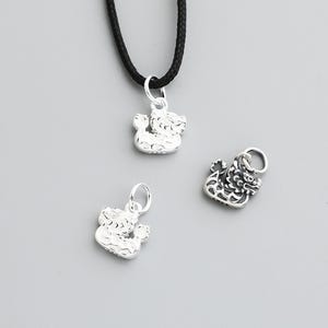 May include: Three silver-colored pendants in the shape of a mythical creature. One pendant is attached to a black cord. The pendants have a detailed, textured design and are displayed against a gray background.