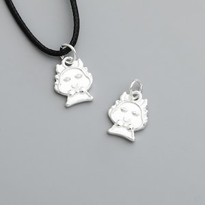 May include: Two silver-colored pendants with a cartoon-like face design. One pendant is attached to a black cord necklace. The pendants have a detailed, slightly raised design. The background is a solid, light gray color.