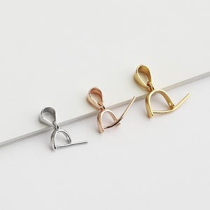 May include: Three metal pendant bails in silver, rose gold, and gold. Each bail has a loop at the top and a horizontal bar extending from the bottom, designed for attaching to jewelry components. The bails are arranged on a white surface.