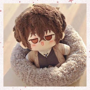 May include: A plush doll of a character with brown hair and eyes, wearing a vest and jacket, resting in a gray, fluffy bed. The doll has a neutral expression with rosy cheeks. The background is a soft, blurred light.