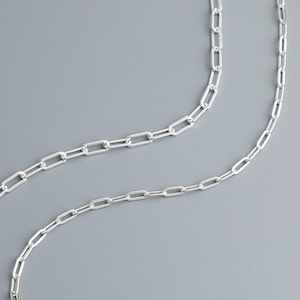 Op de afbeelding: Een zilveren ketting met een delicaat, gekoppeld ontwerp. De ketting is gemaakt van kleine, rechthoekige schakels die aan elkaar zijn verbonden om een ​​continue lus te creëren.