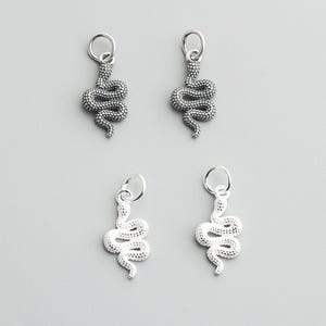 May include: Four silver snake charms with a textured, scaled design. Each charm has a small loop at the top for attaching to a necklace or bracelet. The charms are displayed against a light gray background.