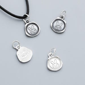 May include: Silver-toned bunny pendant charms. The charms feature a bunny design within a circular frame, with a textured border. One charm is attached to a black cord. The other charms are loose. The charms are small and detailed.