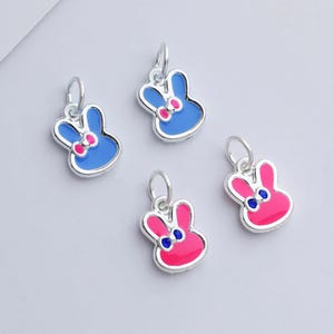 May include: Four silver-toned bunny charms. Two are blue with pink bows, and two are pink with blue accents. Each charm has a small loop at the top for attaching to a necklace or bracelet.