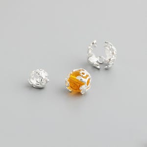 May include: A selection of silver jewellery components. The components include two openwork beads, one with a clear centre and the other with an amber-coloured centre, and a leaf-designed clasp. The components are on a grey background.