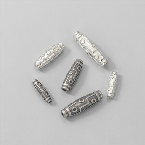 May include: A collection of silver-colored, cylindrical beads with intricate geometric patterns. The beads are arranged on a light gray surface, showcasing their detailed designs and metallic sheen. These beads are suitable for jewelry making.