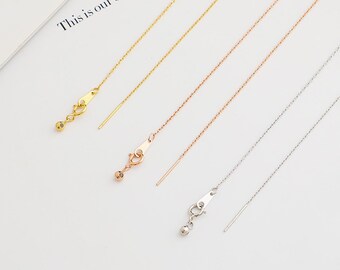 May include: Four delicate chain necklaces in gold, rose gold, and silver tones. Each necklace features a small clasp with a dangling bead and a thin, straight bar. The necklaces are arranged diagonally on a white surface.