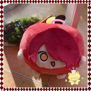 May include: A red plush doll with a cartoon face, orange eyes, and a small mouth. The doll has red hair and small white appendages. A pink ring is attached to the top. The doll is a soft toy.
