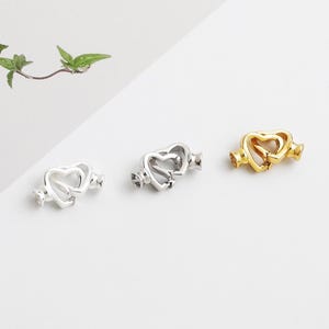 May include: Three heart-shaped jewellery clasps in silver and gold tones. The clasps feature a double-heart design with decorative end pieces. The clasps are displayed on a white surface, with a sprig of green leaves in the background.
