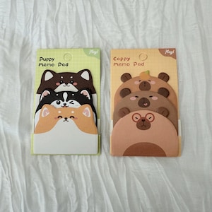 K&ouml;nnte beinhalten: Zwei Notizblock-Sets mit Tiermotiven. Ein Set hat Welpengesichter in Braun, Schwarz und Orange. Das andere Set hat B&auml;rengesichter in verschiedenen Braunt&ouml;nen. Jedes Pad ist mit dem Text "Puppy Memo Pad" und "Cappy Memo Pad" beschriftet.