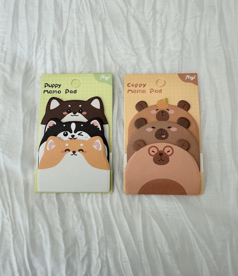 K&ouml;nnte beinhalten: Zwei Notizblock-Sets mit Tiermotiven. Ein Set hat Welpengesichter in Braun, Schwarz und Orange. Das andere Set hat B&auml;rengesichter in verschiedenen Braunt&ouml;nen. Jedes Pad ist mit dem Text "Puppy Memo Pad" und "Cappy Memo Pad" beschriftet.