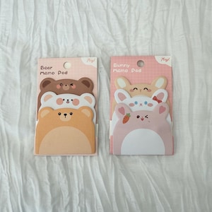 K&ouml;nnte beinhalten: Zwei Notizblock-Sets mit niedlichen Tierdesigns. Ein Set hat b&auml;renf&ouml;rmige Bl&ouml;cke in Braun und Beige, beschriftet mit "Bear Memo Pad". Das andere Set hat hasenf&ouml;rmige Bl&ouml;cke in Rosa und Beige, beschriftet mit "Bunny Memo Pad".