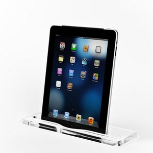 May include: A clear acrylic stand with a black tablet computer resting on it. The tablet screen displays a variety of app icons, including Calendar, Messages, Camera, Videos, and Music. The time is 12:27 AM.