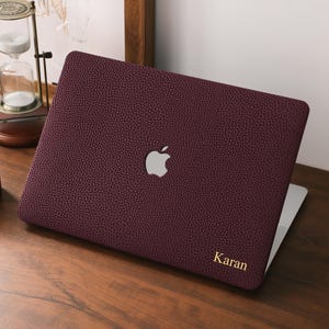 May include: A burgundy laptop case with a textured finish and a silver Apple logo. The name "Karan" is printed in gold on the lower right. The laptop sits on a wooden desk, with an hourglass and a candle visible.