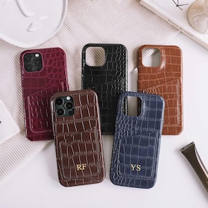 Op de afbeelding: Een verzameling leren telefoonhoesjes in diverse kleuren, waaronder bordeauxrood, zwart, bruin en marineblauw. Elke hoes heeft een krokodillenstructuur en een kaarthouder. Sommige hoesjes zijn voorzien van initialen. Accessoires op een witte ondergrond.