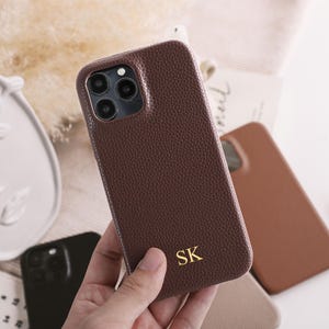 May include: A brown leather phone case with the initials "SK" in gold lettering. The case has a textured surface and a camera cutout. Other phone cases in different colours are visible in the background.