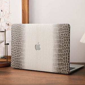 May include: A laptop skin featuring a textured, white and grey crocodile skin pattern. The design includes a central vertical white stripe that transitions into darker grey, scale-like patterns on the sides, with a cutout for the Apple logo.