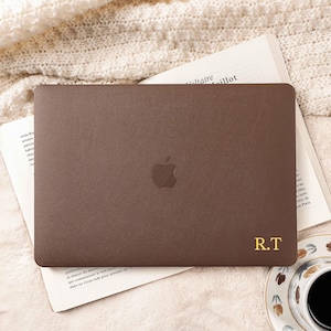 Puede incluir: Una funda marrón para portátil con el logotipo de Apple y las iniciales doradas "R.T." se muestra sobre un libro abierto. Una taza de café y un plato decorativo están en la esquina inferior derecha. La funda está sobre una superficie texturizada color crema.