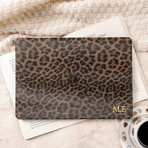 May include: A brown laptop case featuring a leopard print design. Gold initials "M.E" are visible on the bottom right. The case rests on an open book, with a cup of coffee on a saucer in the background, creating a cozy aesthetic.