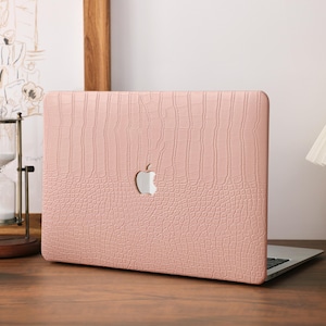 Puede incluir: Una funda rosa para portátil con textura de piel de cocodrilo y un logotipo blanco de Apple. La funda está sobre un escritorio de madera, con un portátil visible. Un reloj de arena, una vela y una lámpara están en el fondo.