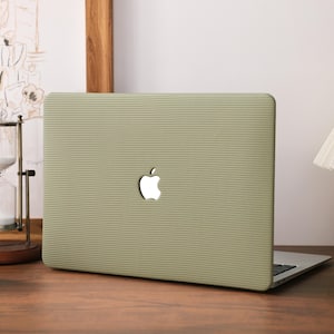 Puede incluir: Un portátil verde salvia con un diseño texturizado y acanalado. El logotipo de Apple es visible en la cubierta del portátil. El portátil está abierto y apoyado sobre una superficie de madera, con una lámpara y un reloj de arena en el fondo.