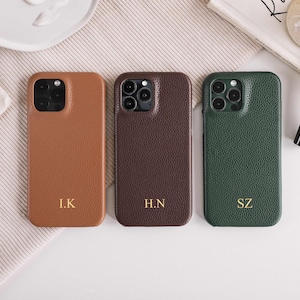 以下が含まれることがあります： ベージュ、ブラウン、グリーンの3色のレザー製携帯電話ケース。各ケースはテクスチャ加工された表面と金色のイニシャルが付いています。ケースは、他のアクセサリーと一緒にニュートラルな表面に配置されています。