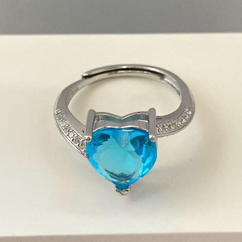 K&ouml;nnte beinhalten: Ein silberner Ring mit einem gro&szlig;en herzf&ouml;rmigen blauen Edelstein und kleineren klaren Edelsteinen um das Band.