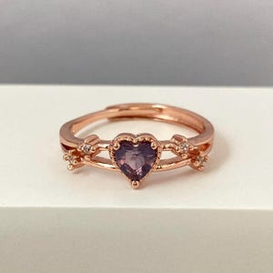 以下が含まれることがあります： ハート型の紫色の宝石と小さな透明な石が特徴の、繊細なデザインのローズゴールドのリングです。