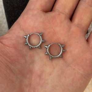 May include: A pair of silver-toned hoop earrings featuring small, evenly spaced metal spikes protruding from the outer edge of each circular band. The earrings have a polished metallic finish and a hinged closure mechanism visible on the hoops.